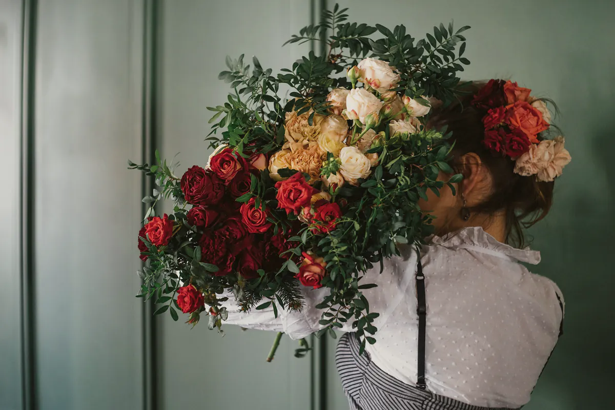 A florist in an apron carrying an armful of red, apricot and cream roses with trailing greenery past a green panelled wall