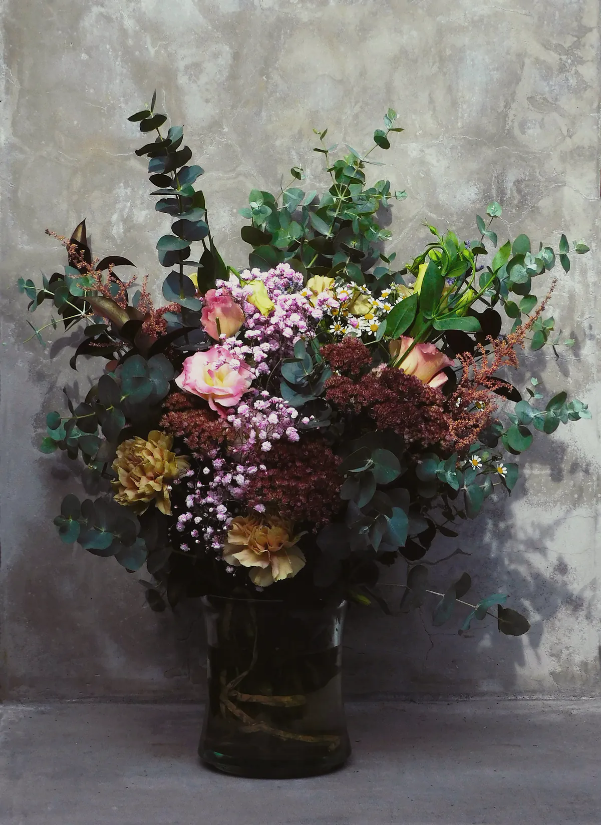 A tall, overflowing arrangement of eucalyptus, roses and dried grasses in a smoky glass vase