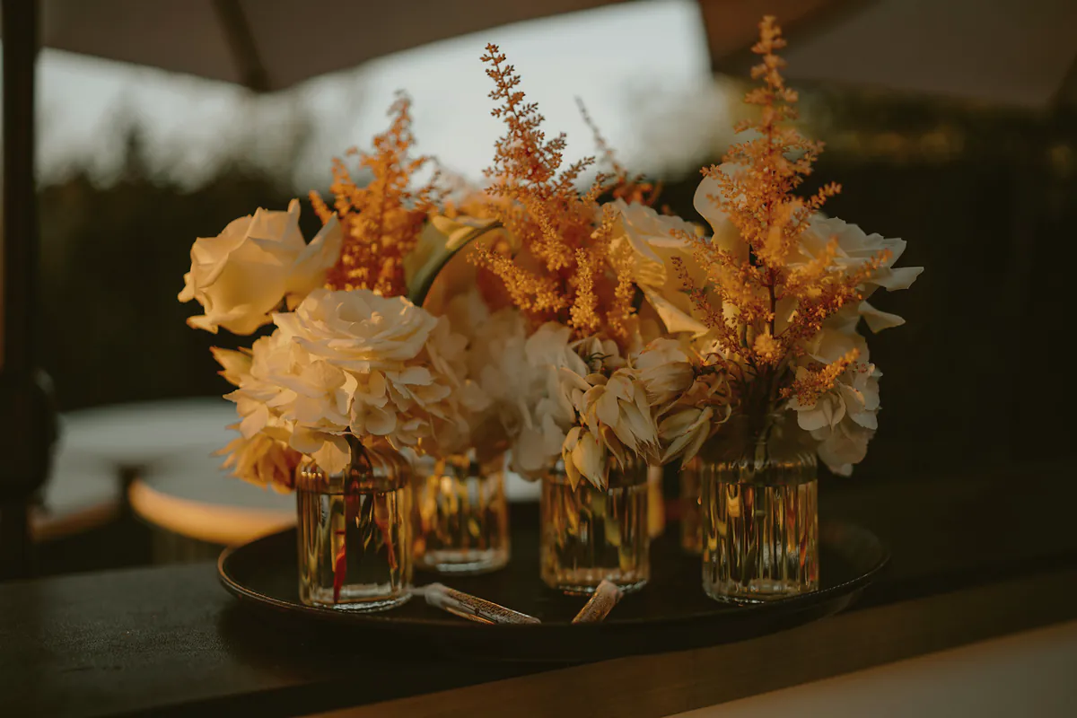 Small ribbed glass vases of cream roses, hydrangea and apricot astilbe on a dark tray at dusk