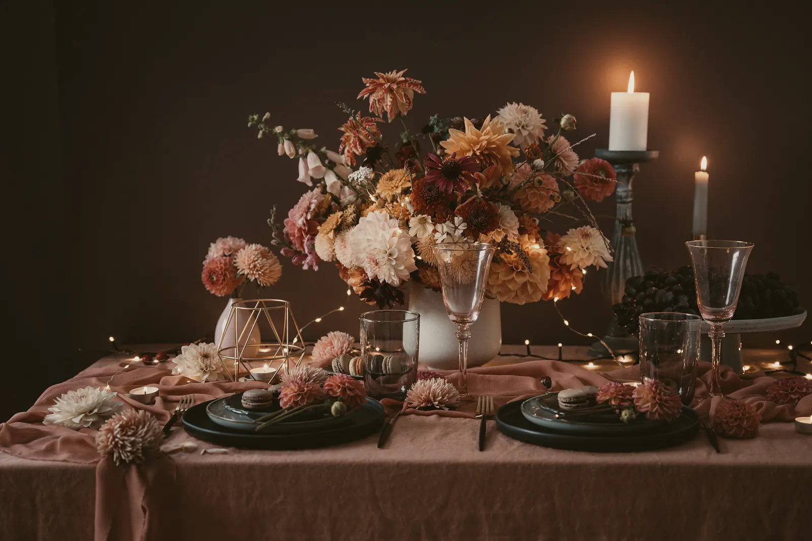 A candlelit dinner table with a tall arrangement of dahlias, foxgloves and cosmos on a dusty pink linen cloth
