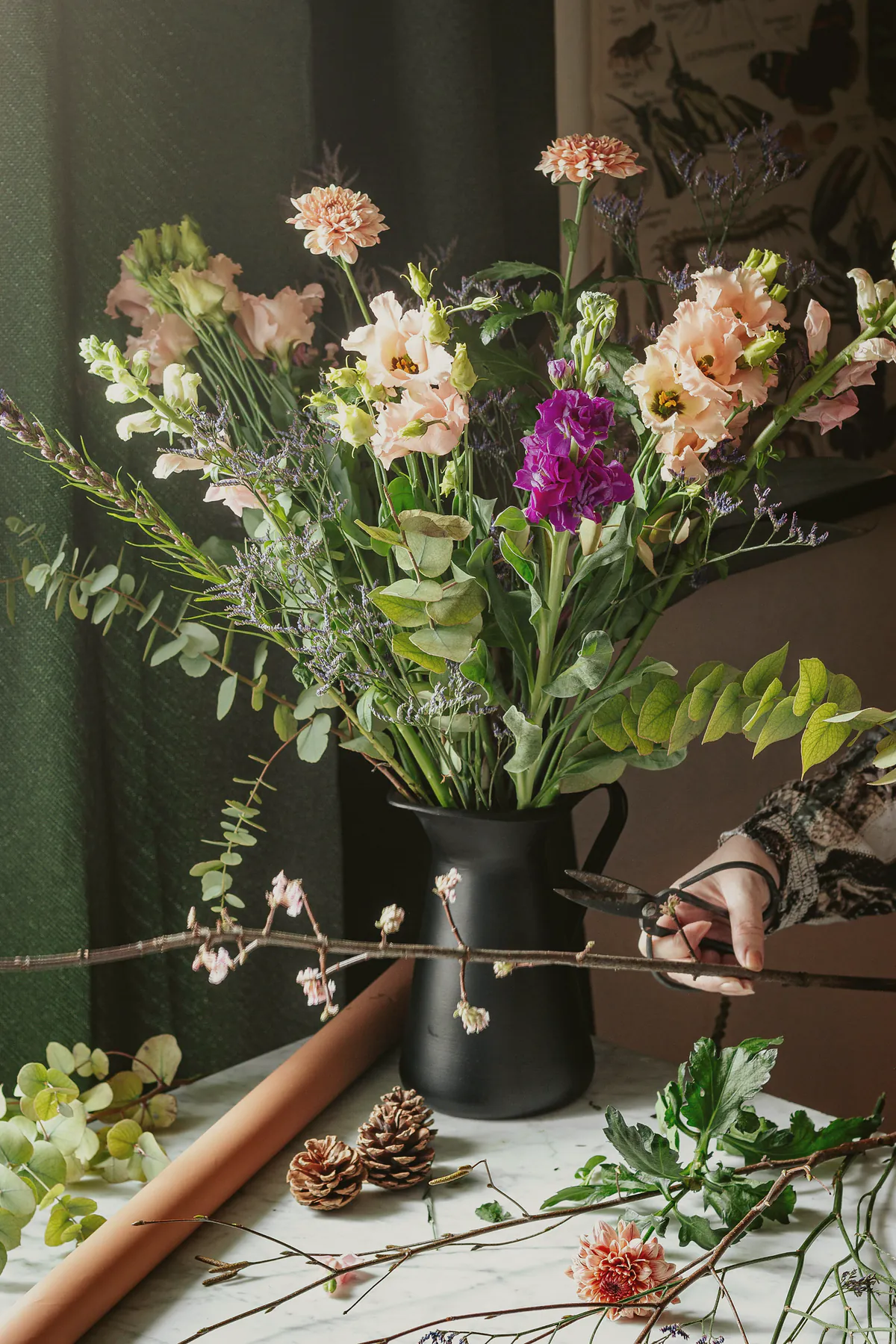 A black jug of peach and magenta flowers with bare blossom branches on a white bench