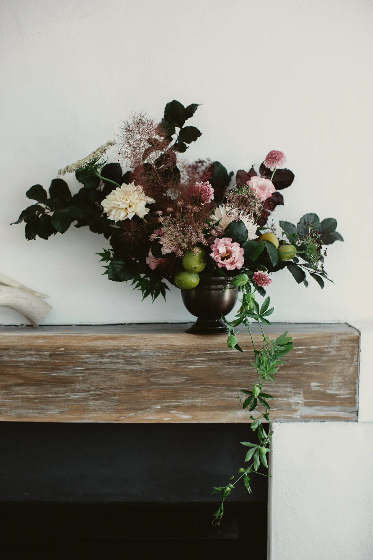 A dark footed urn of blush roses, foliage and trailing vines on a timber mantelpiece