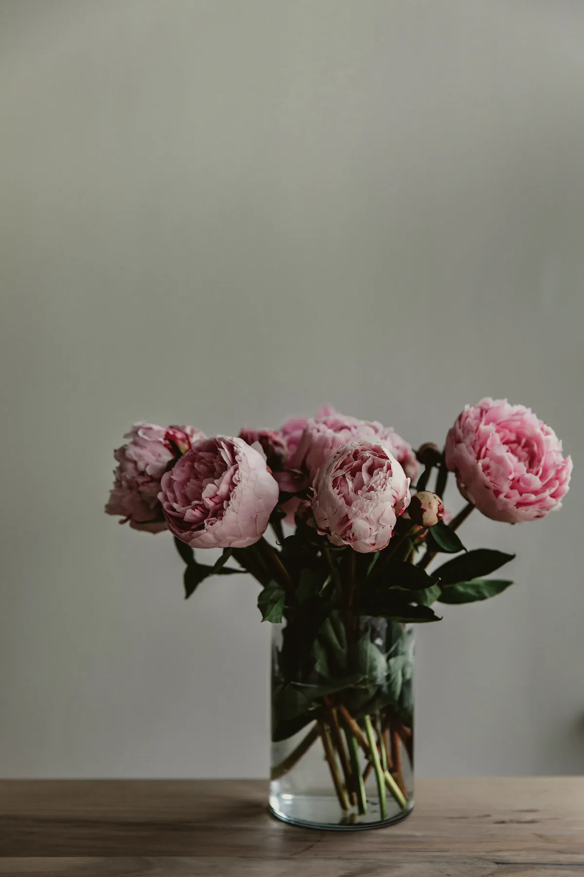 Five pink peonies in a simple glass jar on a timber table
