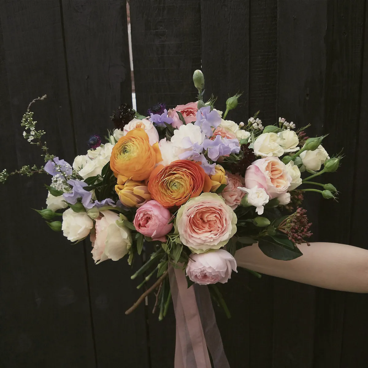 A hand holding out a bouquet of orange ranunculus, peach garden roses, lilac sweet peas and white roses against black timber