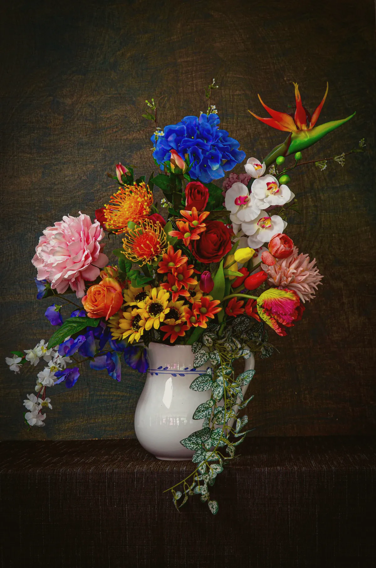 A jug of dahlias, blue delphinium, orchids and strelitzia lit like a still life painting
