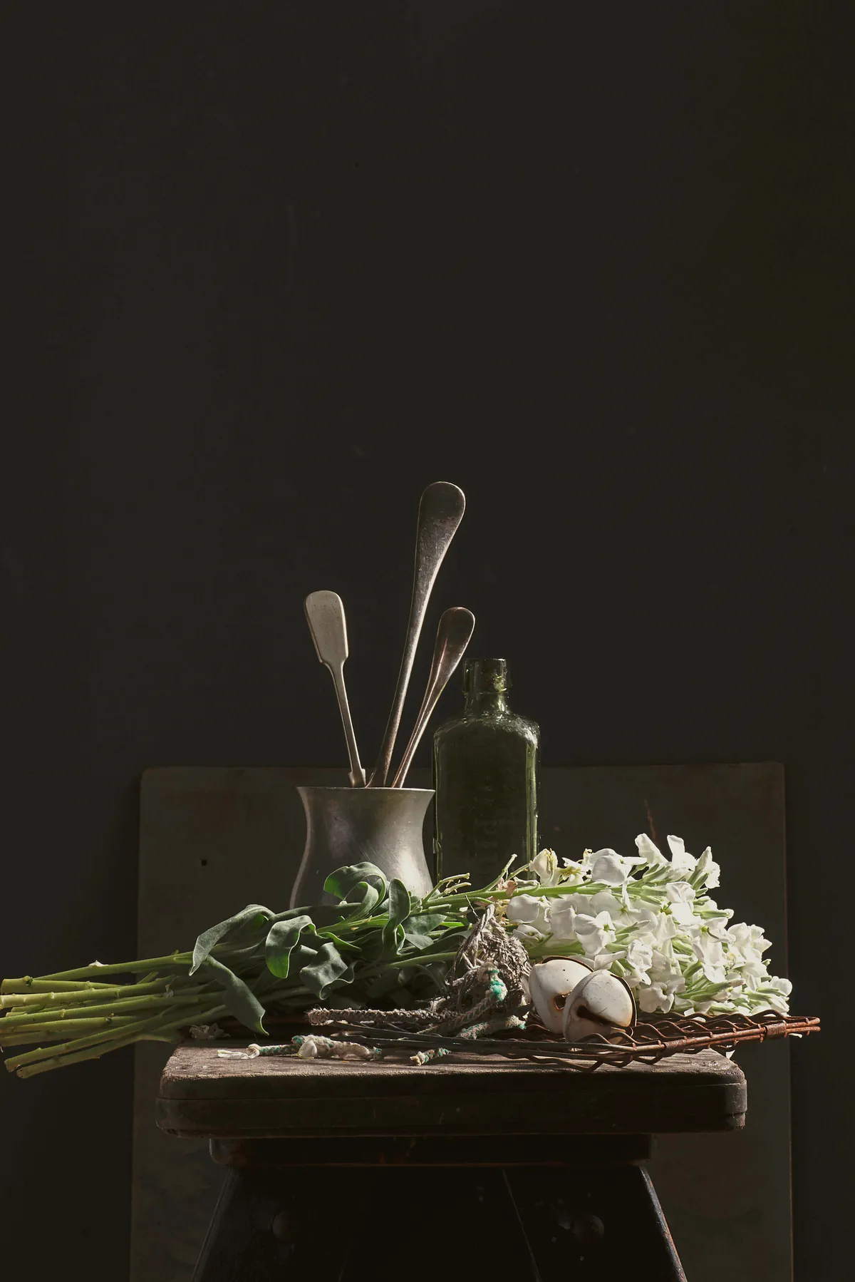 White stock and tulip leaves laid on an old stool beside a pewter jug of silver spoons and a green glass bottle