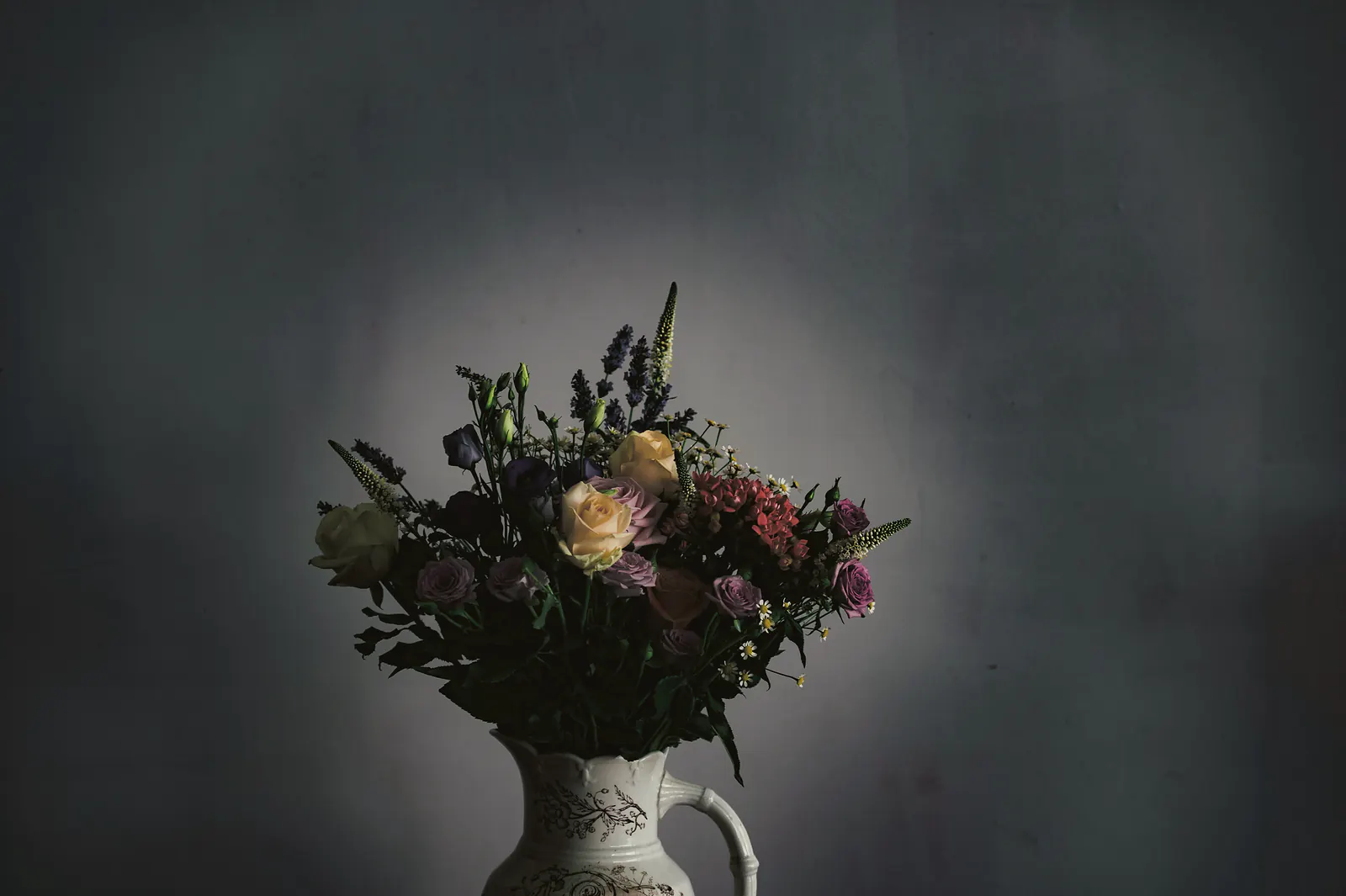 A loose bouquet of roses, lisianthus, lavender and sweet William in an old transfer printed jug, lit by a single soft light