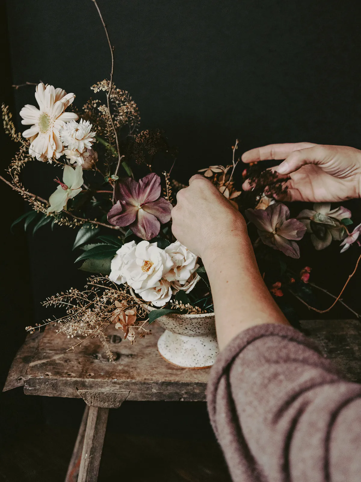 A florist’s hands working hellebores, garden roses, a pale gerbera and dried grasses into a speckled bowl on an old timber stool