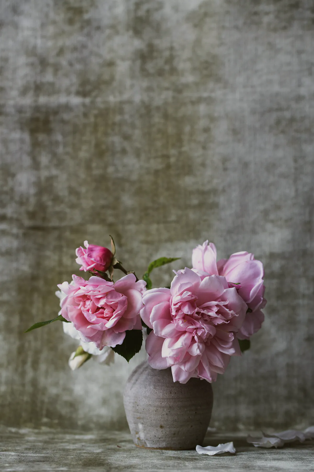 Pink peonies spilling from a small grey stoneware pot against a weathered plaster wall