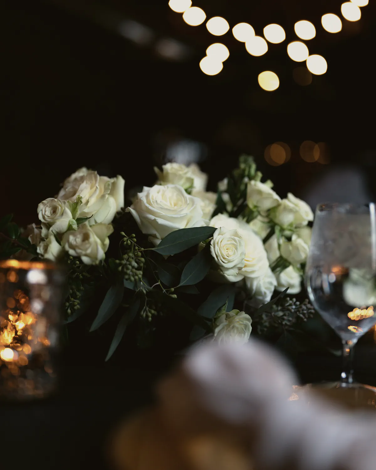 White garden roses and spray roses with eucalyptus on a candlelit reception table, a water glass beside them and warm lights overhead