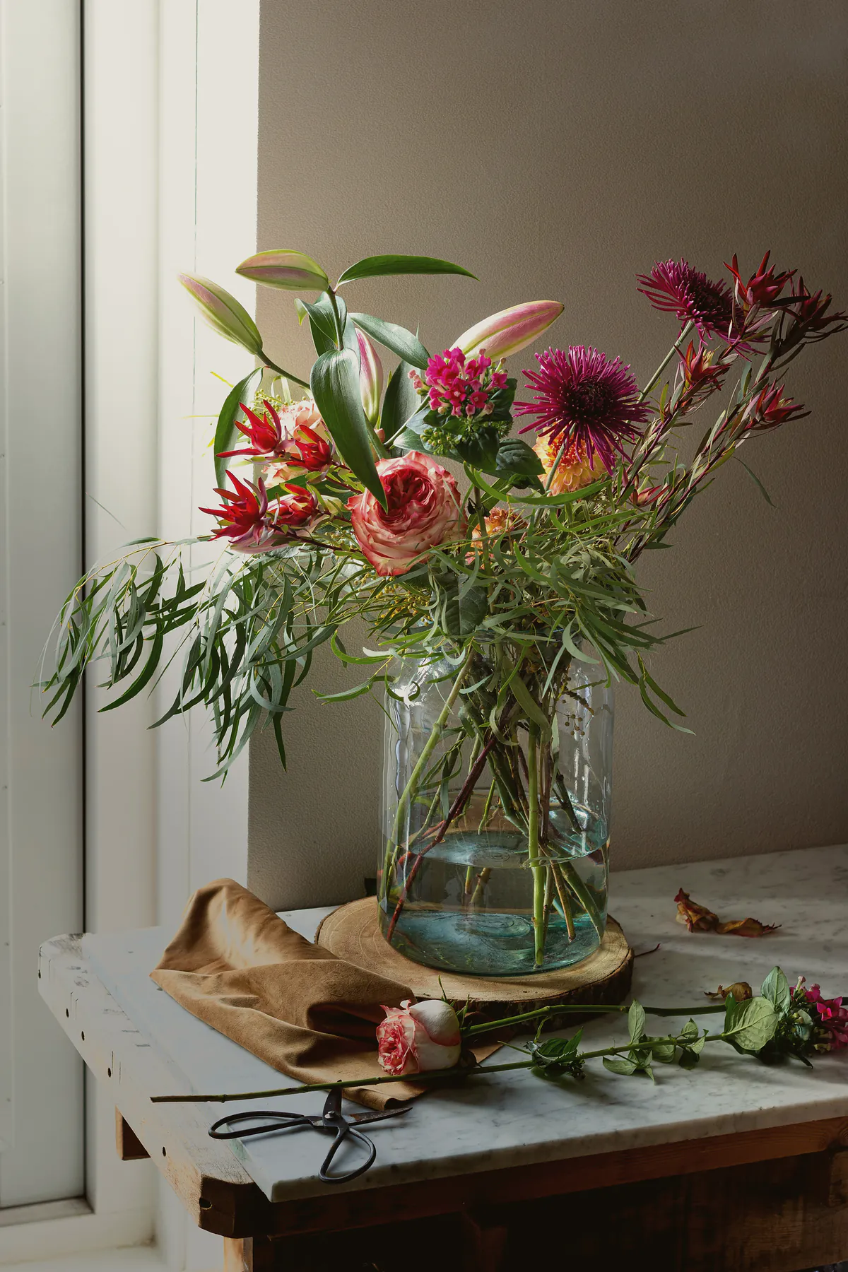 A glass jar of dahlias, lilies and willowy greenery by a sunny window, with scissors on the bench